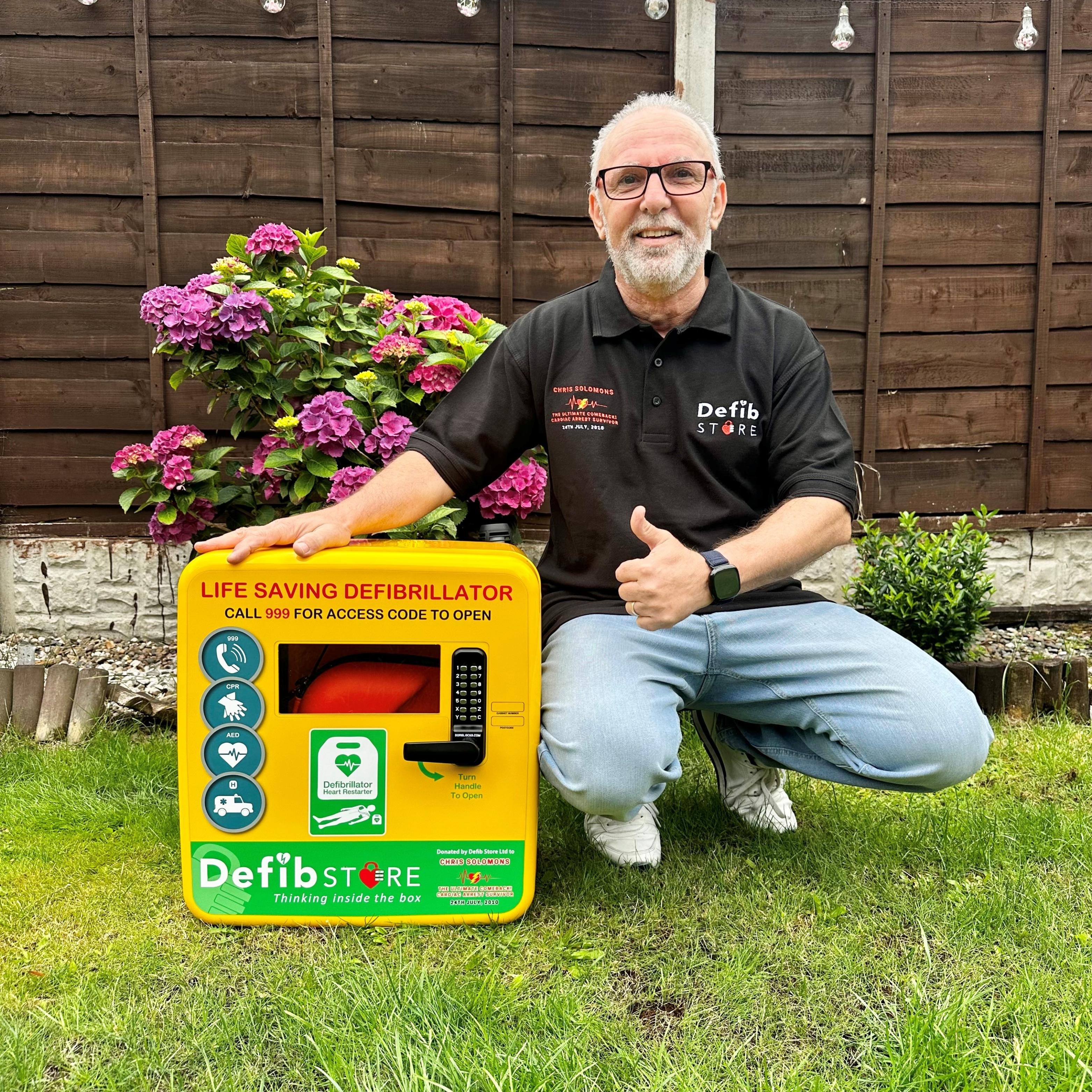 Chris Solomons, cardiac arrest survivor and Defibstore brand ambassador, standing with a Defibstore defibrillator cabinet.